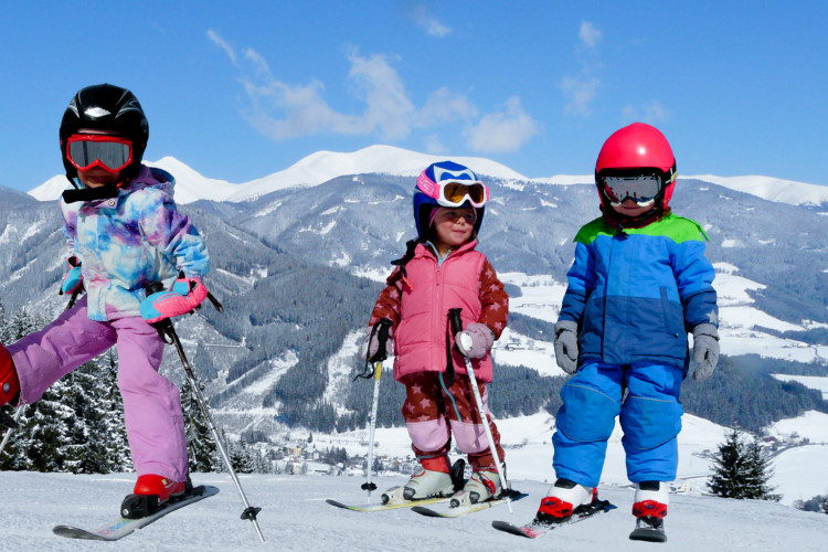 Drei Kinder in bunter Skibekleidung stehen lachend auf Skiern im Schnee, mit verschneiten Bergen und blauem Himmel im Hintergrund.