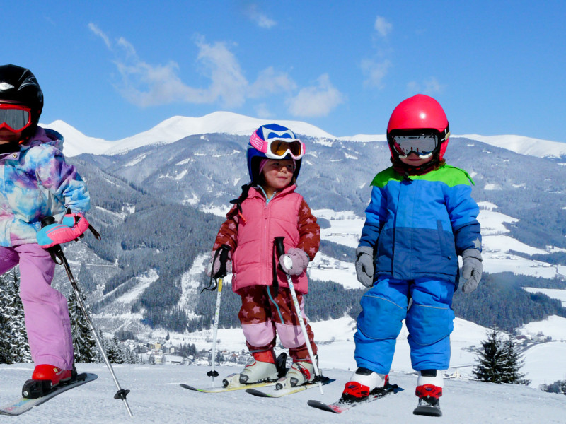 Drei Kinder in bunter Skibekleidung stehen lachend auf Skiern im Schnee, mit verschneiten Bergen und blauem Himmel im Hintergrund.