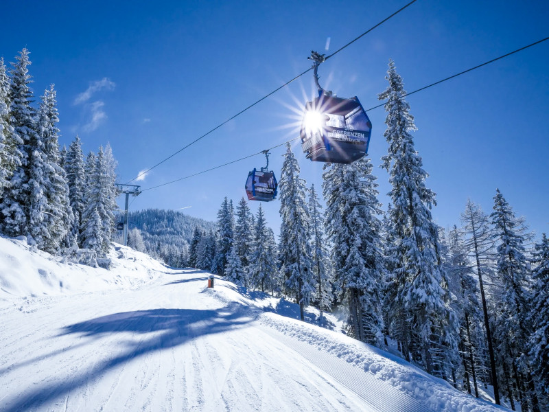 Seilbahn-Gondeln fahren über eine verschneite Skipiste durch winterliche Tannenwälder bei strahlendem Sonnenschein und blauem Himmel.