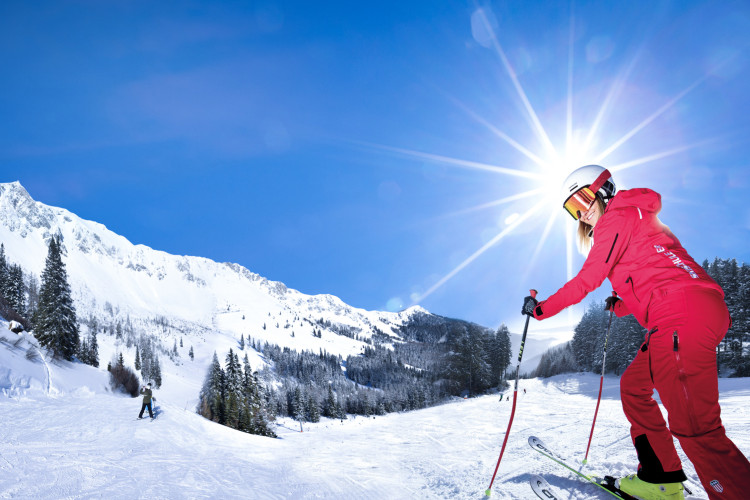 Skifahrerin in roter Skibekleidung fährt lachend eine sonnige Piste hinunter, im Hintergrund verschneite Berge, Tannen und strahlender Sonnenschein.