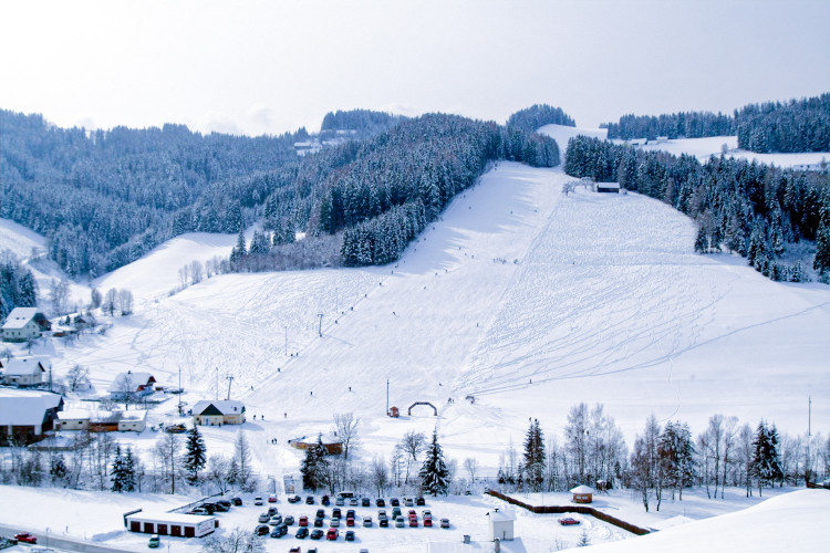Panoramaaufnahme des verschneiten Kleinlobming Skigebiets mit Skipisten, Skiliften, umgeben von Wäldern und einem Tal mit Häusern und Parkplatz im Vordergrund.