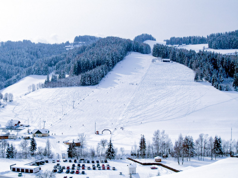 Panoramaaufnahme des verschneiten Kleinlobming Skigebiets mit Skipisten, Skiliften, umgeben von Wäldern und einem Tal mit Häusern und Parkplatz im Vordergrund.