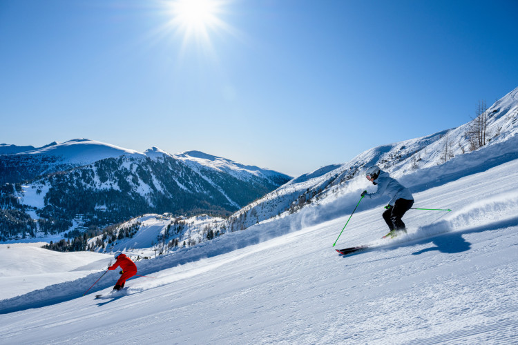 Zwei Skifahrer fahren bei Sonnenschein eine verschneite Piste hinunter, umgeben von beeindruckenden Berggipfeln und tiefblauem Himmel.