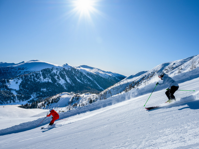 Zwei Skifahrer fahren bei Sonnenschein eine verschneite Piste hinunter, umgeben von beeindruckenden Berggipfeln und tiefblauem Himmel.
