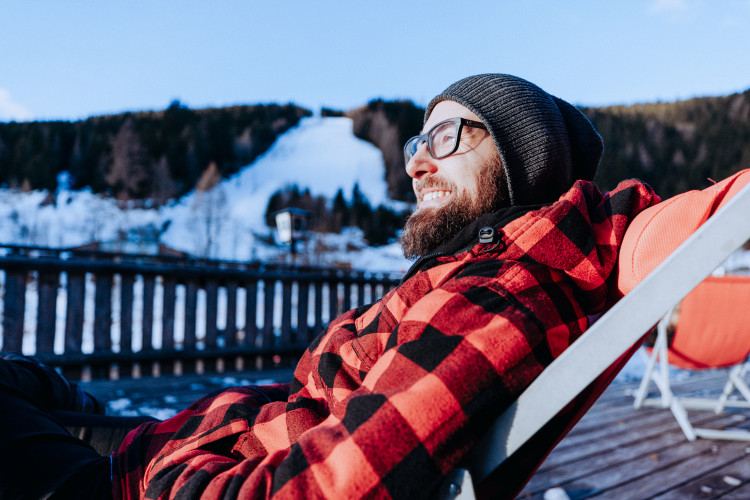 Mann mit Bart, Brille und Mütze entspannt in einem Liegestuhl auf einer Sonnenterrasse. Im Hintergrund ist unscharf eine verschneite Piste zu sehen.