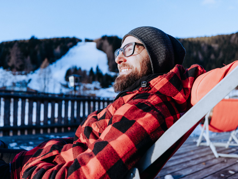 Mann mit Bart, Brille und Mütze entspannt in einem Liegestuhl auf einer Sonnenterrasse. Im Hintergrund ist unscharf eine verschneite Piste zu sehen.