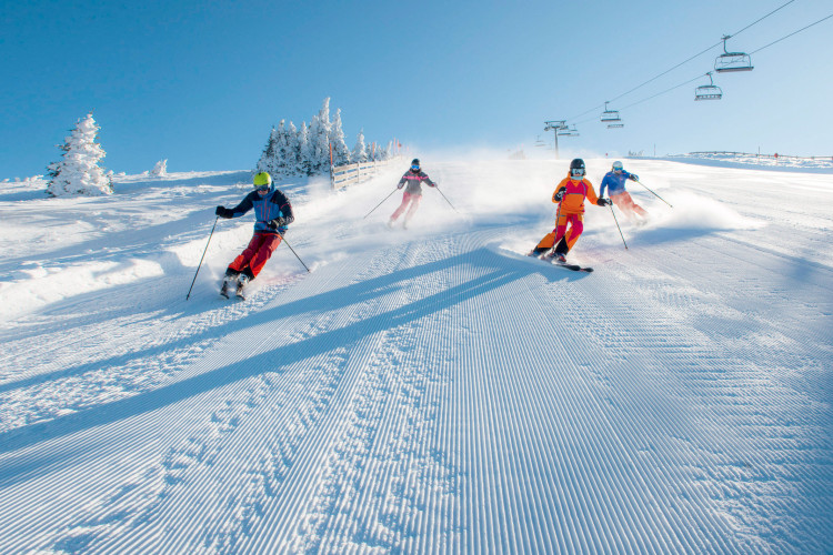 Gruppe von Skifahrern fährt schwungvoll eine frisch präparierte Skipiste hinunter, umgeben von verschneiten Bäumen und Sesselliften bei strahlend blauem Himmel.