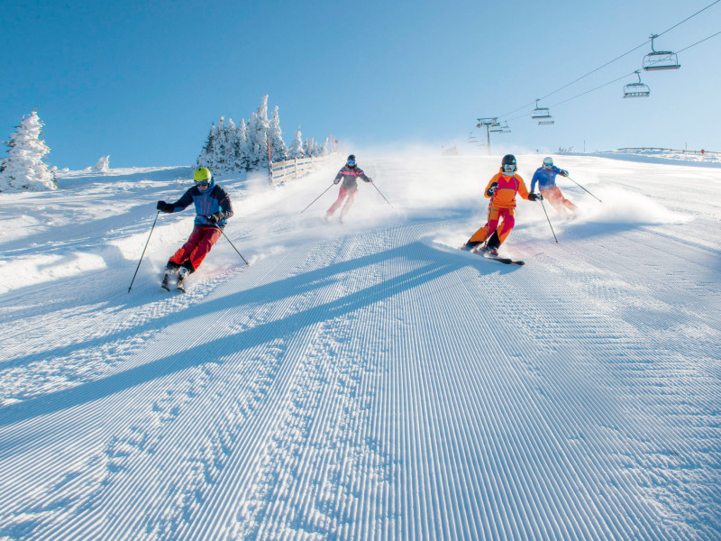 Gruppe von Skifahrern fährt schwungvoll eine frisch präparierte Skipiste hinunter, umgeben von verschneiten Bäumen und Sesselliften bei strahlend blauem Himmel.