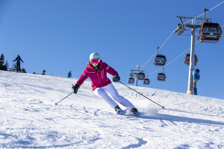 Skifahrerin in pinker Jacke und weißer Hose fährt schwungvoll eine sonnige Piste hinunter, im Hintergrund ist der Sessellift am Kreischberg zu sehen.