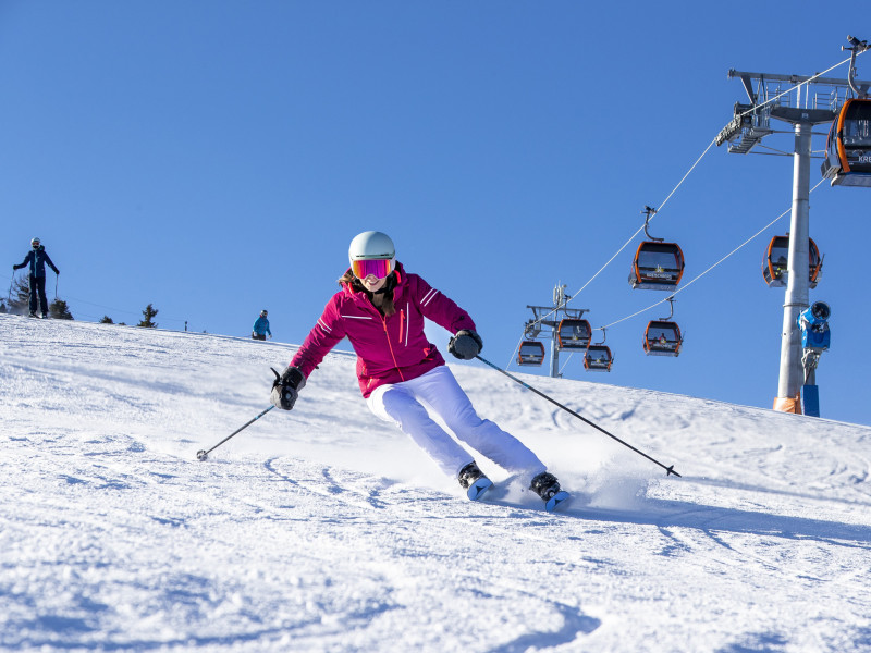 Skifahrerin in pinker Jacke und weißer Hose fährt schwungvoll eine sonnige Piste hinunter, im Hintergrund ist der Sessellift am Kreischberg zu sehen.