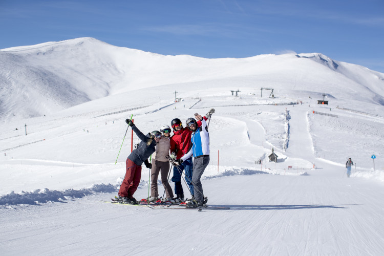 Gruppe von vier Skifahrern posiert lachend auf der Skipiste mit schneebedeckten Bergen im Hintergrund bei sonnigem Winterwetter.