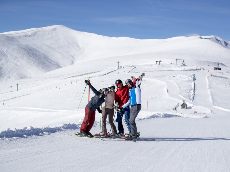 Gruppe von vier Skifahrern posiert lachend auf der Skipiste mit schneebedeckten Bergen im Hintergrund bei sonnigem Winterwetter.