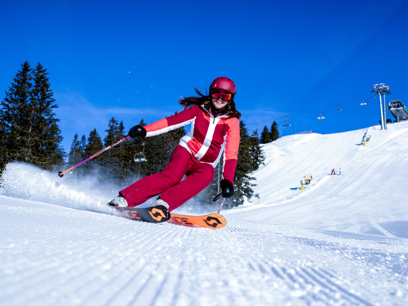 Frau in roter Skibekleidung fährt dynamisch eine sonnige Skipiste hinunter.