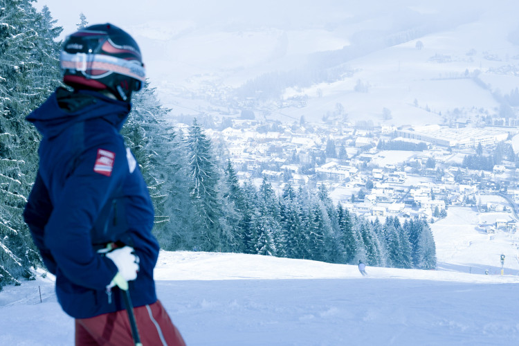 Skifahrer mit Helm und Skistöcken blickt von einer verschneiten Piste auf ein winterliches Tal der Stadt obdach im Hintergrund, umgeben von verschneiten Wäldern.